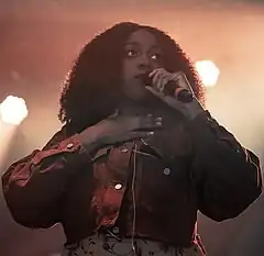 Noname onstage smiling, holding a microphone