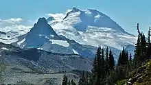 A prominent, glaciated mountain overlooking a rocky peak on the left and a forested slope on the right.