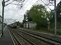 A train approaches the station. The former platform is on the right. The new platform is on the other side of the level crossing.