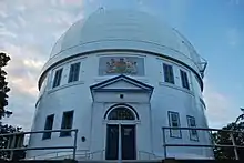 Colour photograph of the two-storey building, clad in painted white metal panels, with a projecting entrance portico consisting of blue double doors. The Dominion of Canada coat of arms is seen above the door.