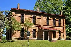 A two-story brick building with three chimneys on top of a grassy hill. There are bars on the long, arched windows. There are trees on either side and a historical marker is visible in the foreground.