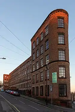 Slim multi storey dark red brick building on the narrow junction of two roads against a pale blue sky