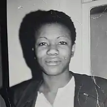 Black and white head and shoulder photograph of a black woman with a close-cropped hair style in a jacket and v-necked white blouse. A shadow frames the right side of her face and a narrow window is to her left.