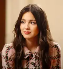 A mid-shot of a young woman with long, curly brown hair, wearing a fur blazer-style jacket. She is looking towards the left of the camera and smiling.