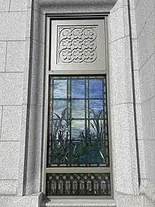 Stained glass depicting various water elements and bullrushes, against a blue sky background. A metal frame outlines the glass and above the stained glass depicts a pattern of nine cherry blossoms. Masonry from the temple walls hold the window in place.