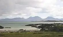 A small sandy beach lies beyond a grassy foreshore. In the distance, the outline of a range of brown and grey hills is visible under cloudy skies.