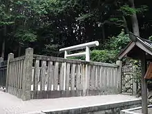 White torii gate behind a concrete fence in front of woods.