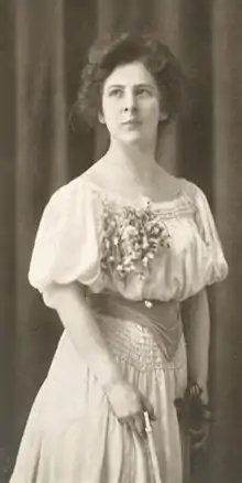 A young white woman in a white dress, standing; in her hands are the bow and neck of a violin.