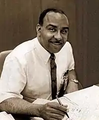 portrait seated at his desk in shirtsleeves and tie with pen in his hand