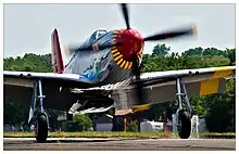 A single nosed-propeller plane is on the ground on its wheels with the propeller in motion.  The plane is viewed from the front, but the red nose faces slightly to the right.  The plane has black propellers.  Parts of the wings and propellers are crpped from view.