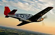 A single nosed-propeller aircraft is in mid-flight above land.  The red nose faces right and the underside of the aircraft is slightly in view.  The aircraft has black propellers, gray base paint, and black letters reading: "A42".  The "4" and "2" are separated by a United States roundel in black with a central white star.  The roundel is also visible on the tops of the wings.  The full length of the right side of the aircraft is visible.