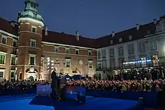 A man in a suit stands at a podium; an audience of people is assembled in a U around him