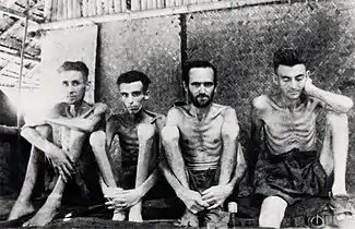 Four malnourished shirtless men sit against a wall