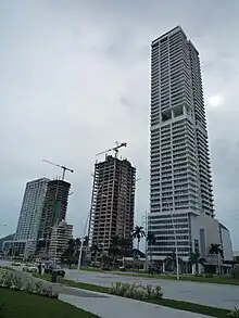 Two tall buildings, with construction cranes on top, next to much taller skyscraper