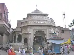 An elaborate, carved entrance to a Hindu temple whose canopy is visible at the top of the image. The entrance section is polygonal with arches and there is a stone staircase leading into the grey/cream coloured structure. Several pilgrims are seen in the foreground, as is a stall.