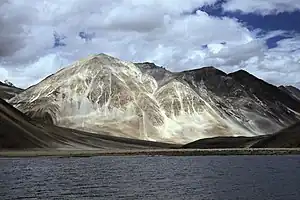 Daytime view of a large body of water standing before a prominent peak, which communicates with several others partly out of view and behind. A gravel beach at the far end of the lake gives way to steep slopes leading up to the peaks; The mountains lack trees. Patchy snowcover defines their recesses, and whitish vein-like streaks extend up from the base of the largest.