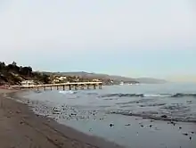 Photograph of a beach. Image of a beach. In it, a pier and several houses can be appreciated.