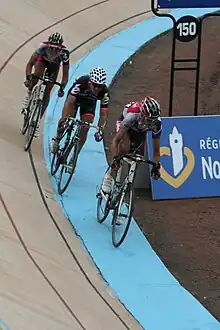 Three cyclists in a line on a racing track. The first and the third in line wear identical red, black, and white jerseys, while the cyclist in the middle wears a Cervélo jersey.