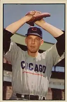 A man in a white baseball uniform with dark sleeves and a dark cap holds his hands together over his head in his glove