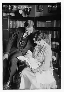 A white man in a suit and a white woman in a dress; the woman is holding a baby, presumably their daughter; both adults are looking at the baby. They are posed in front of a bookcase.