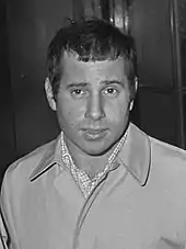 An image of a man in a black and white filter dressed in a coat and dress shirt looking towards the camera.