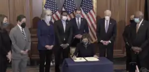 Speaker of the House Nancy Pelosi seated at a table and surrounded by public officials. She is signing the second impeachment of Trump.