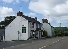 white-painted roadside pub with white-painted house and woods beyond under partly cloudy and blue sky