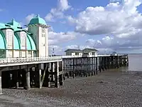 A pier beneath a cloudy sky, with a large white and green building and some smaller huts on it