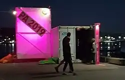 A large pink light box with a diagonal banner PE2019 next to an open shipping container and people walking past - image taken at night time.