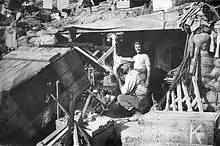 A man in wearing a military-style cap kneels behind rifle that has been modified to be fired from behind the cover of a makeshift breastworks