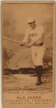 A sepia-toned baseball card image of a man wearing an old-style white baseball uniform and pillbox cap holding a baseball bat extended with both hands