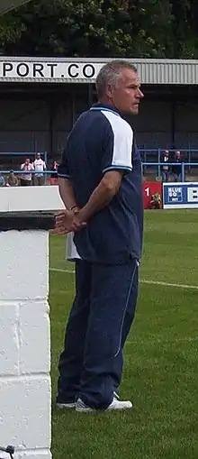 A middle-aged man with greying hair, wearing a dark blue T-shirt and matching jogging bottoms, standing near the touchline of a sports pitch. Some spectators are visible in the background.