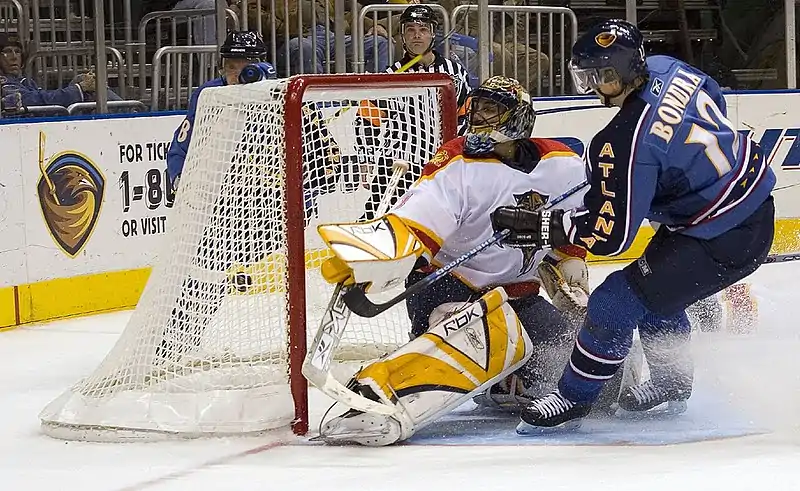 An ice hockey player wearing a blue jersey following through on a shot against a goaltender wearing a white jersey from close proximity. The goaltender's left blocker and pad are outstretched as he watches the puck go in the net behind him.