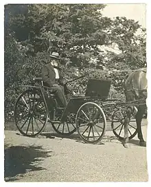 A black and white photograph of an older man wearing a white hat seated in an open carriage.