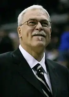 A man with white hair, wearing a black suit, white shirt and tie, at a basketball game.