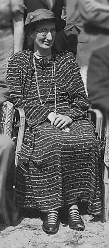 A 40-year-old woman sitting in a wicker chair wearing a long, dark dress. She also wears spectacles and a sunhat.