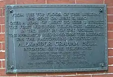 An image of darkened brass historical plaque with a streak of green corrosion running down it, mounted on the exterior side of a brick building.