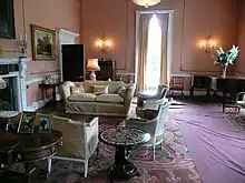 Drawing room with high ceilings and tall window having biscuit coloured walls with white raised mouldings, soft furnishings and piano in corner