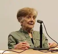 A color photograph of a woman seated at a table behind a microphone