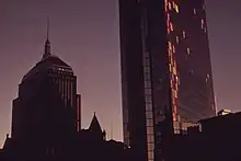 Two dark buildings rise into the early evening sky. The tower on the right is spotted with plywood on its side.