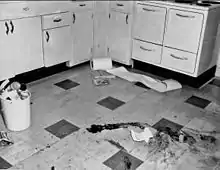 A black and white image of a checkered tiled floor with a right-angled row of light-colored cabinets at the rear. On the floor in the foreground are several large dark smears, with a small piece of paper on the ground nearby. At the left is a small white plastic bucket filled to over the top with trash; some cans and a telephone handset are visible. In the rear, near the cabinets, is a roll of paper with part of it spilling off to the right; underneath it are what appear to be two books