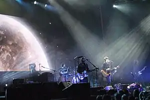 A stage shot of six men performing: From left, a man seated at keyboards with a microphone, the drummer behind his kit with a drumstick raised in his right hand, a guitarist singing into a microphone, another singer-guitarist highlighted by overhead lighting, a man playing a bass guitar who is mostly obscured behind the lead singer, and the last man at right who is also playing a guitar. In front of the stage are cameramen and audience members. Above is further stage lighting with, at left, a large projected image of the top part of the moon.