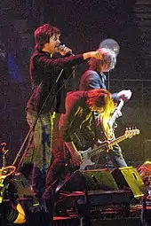 Frontline of Powderfinger are shown in side profile across a stage, with Fanning closest to viewer. He is pointing into the audience with his right fore-finger, while singing into the microphone held in his left hand. Just beyond him, Collins is stooped low over his bass guitar with his long hair fallen forward. Haug is further along and plays his guitar while standing more upright, his shoulder length hair partly obscures his face. Stage equipment is in front of the three men.