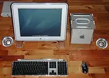 The G4 Cube sits besides a large, flatscreen monitor in matching grey. Flanking the monitor and Cube are round speakers clad in translucent plastic, and a keyboard and mouse.