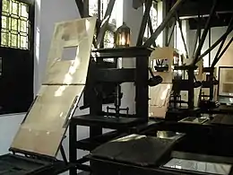 A row of printing presses in a room with lamps and windows on the wall behind them