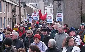 A group of more than 30 protestors in winter clothing carrying protest placards in opposition to the nuclear power station. Located in a village street of terraced houses