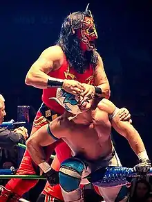 A picture of Delta sitting on the top rope of a wrestling ring, with Psicosis standing behind him tearing at Delta's mask