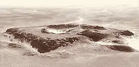 Black and white aerial photograph of the ruins of a square-shaped monument with a depression in the center