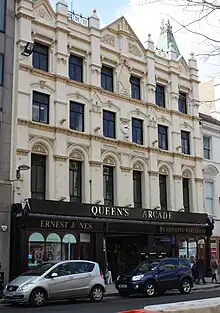 An ornate Victorian 4-storey building, painted pastel yellow, featuring muted copper spires on the roof, a small white clockface in the centre of the 2nd floor, and ground floor shopping units framed in black, either side of the arched entrance to an arcade. On the third floor is an Art-Deco-influenced monogram with the letters "A.R.".