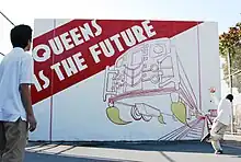 Two children playing handball in front of the Queens Is The Future mural, painted on a handball court in Jackson Heights, Queens. The children are middle-school aged and wear school uniforms. One has just hit the balll. The other waits to receive it. The ball is in mid-air.